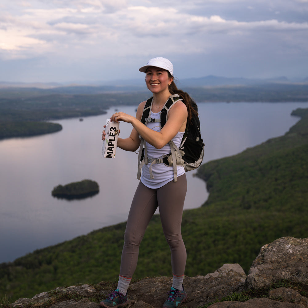 A girl enjoying her plant-based maple water on top of a mountain with a beautiful view on a lake.