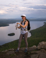 A girl enjoying her plant-based maple water on top of a mountain with a beautiful view on a lake.
