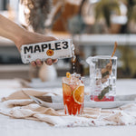 Pouring Maple 3 peach and mango sparkling water into an orange cocktail.